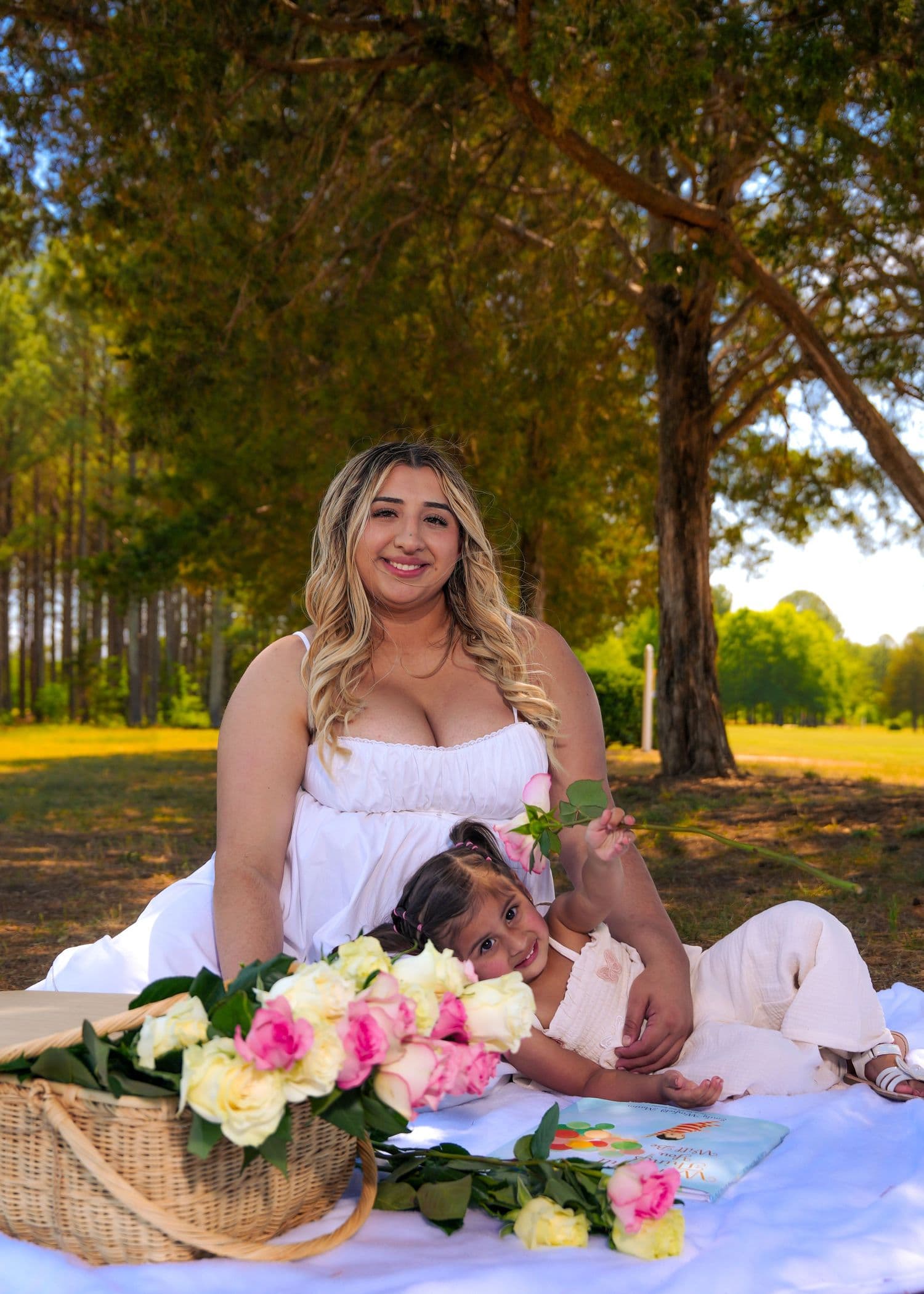 Mother and daughter on a white blanket with a basket of roses under tall trees
