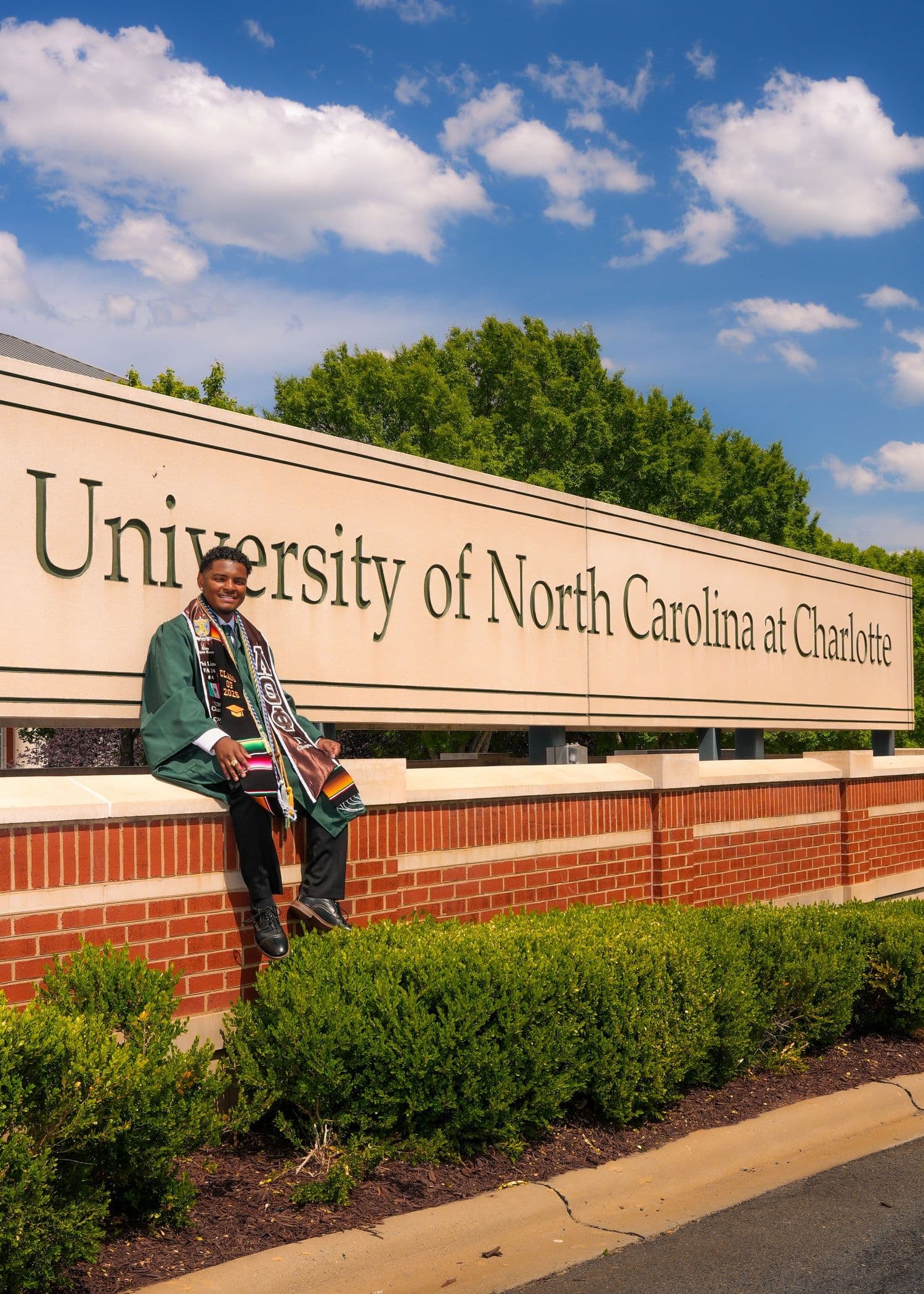Graduate sitting on the University of North Carolina at Charlotte entrance sign under a blue sky