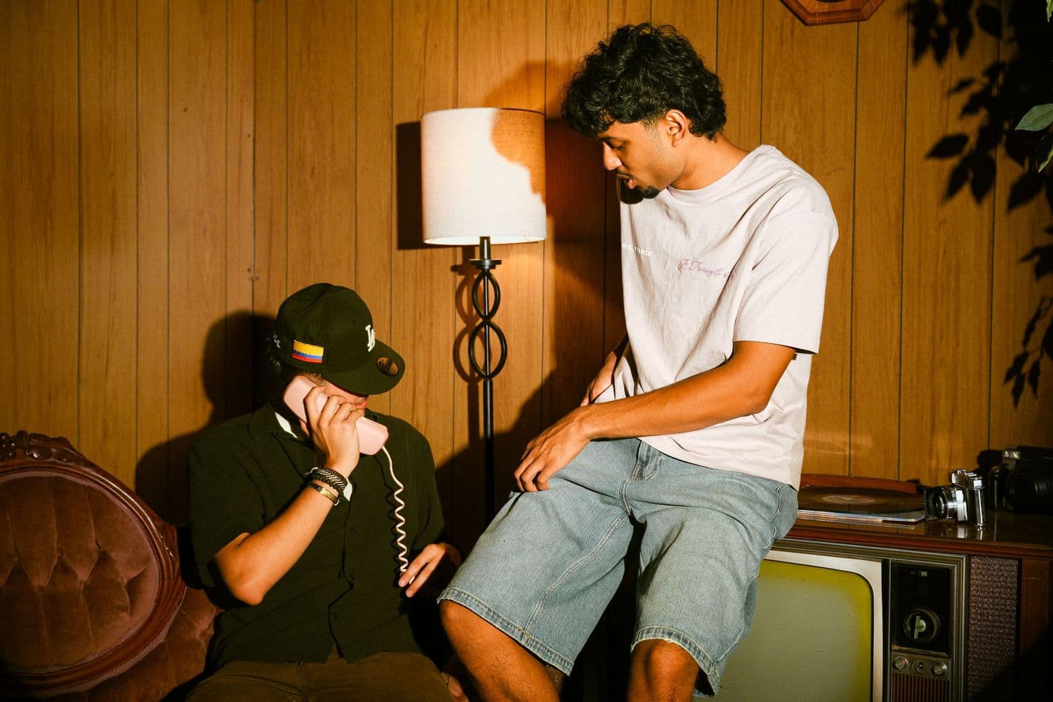 Two friends in a wood-paneled retro room, one on a corded phone, lit with direct flash
