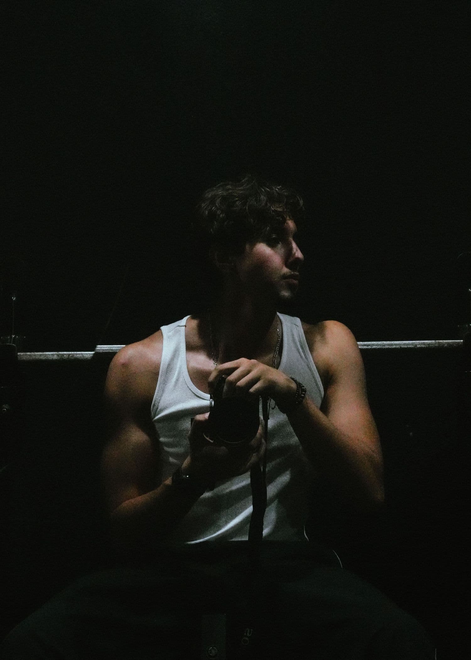 Athlete in a white tank top gripping a bar in a dark, moody gym