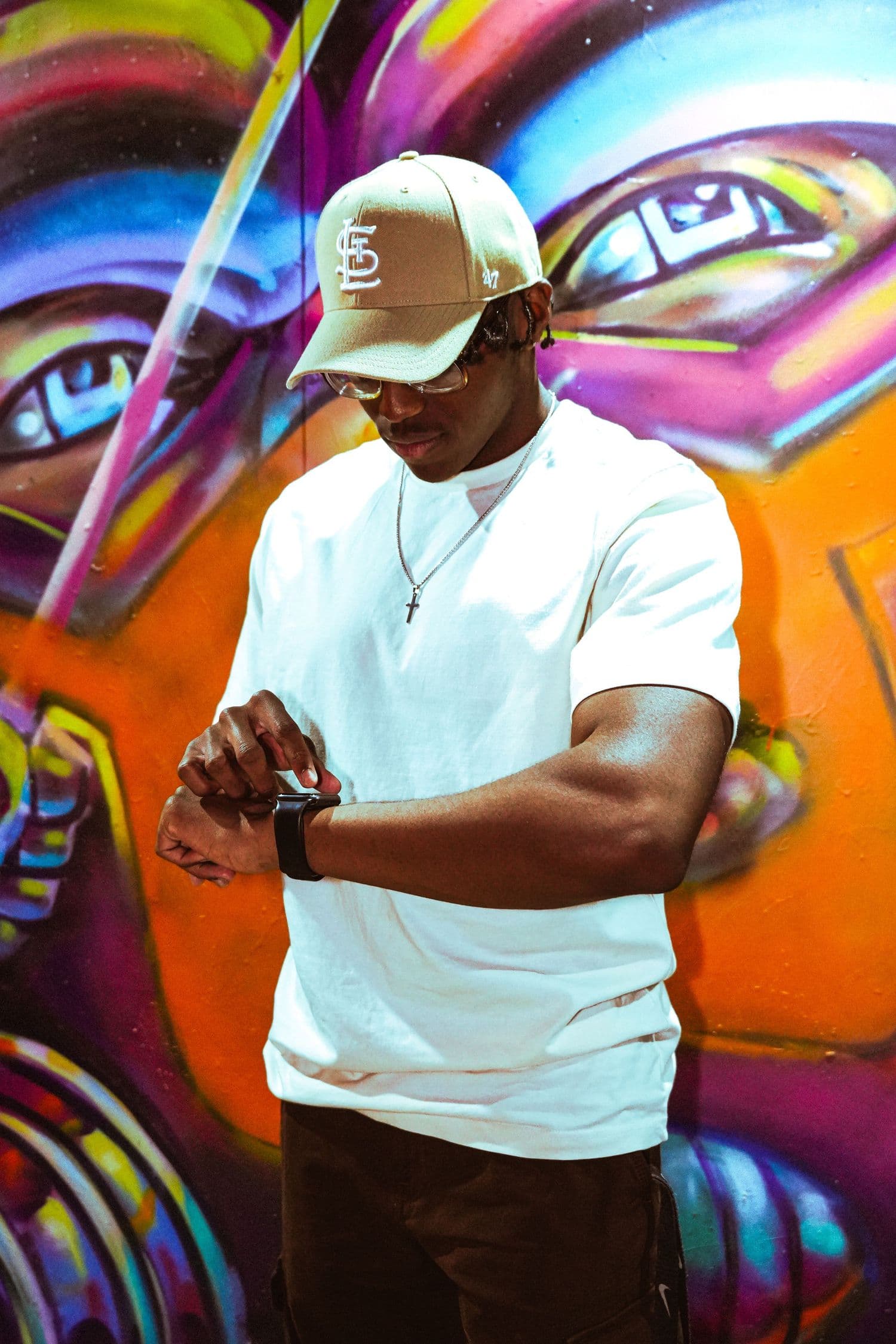 Portrait in front of a colorful street mural: white tee, tan cap, checking a watch